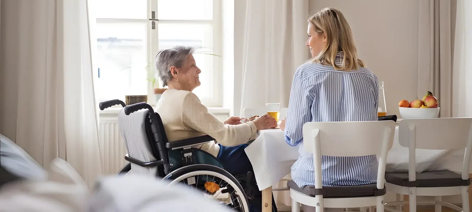 Seniorin im Rollstuhl und eine jüngere Frau sitzen im Gespräch am Tisch beim Kennenlernen